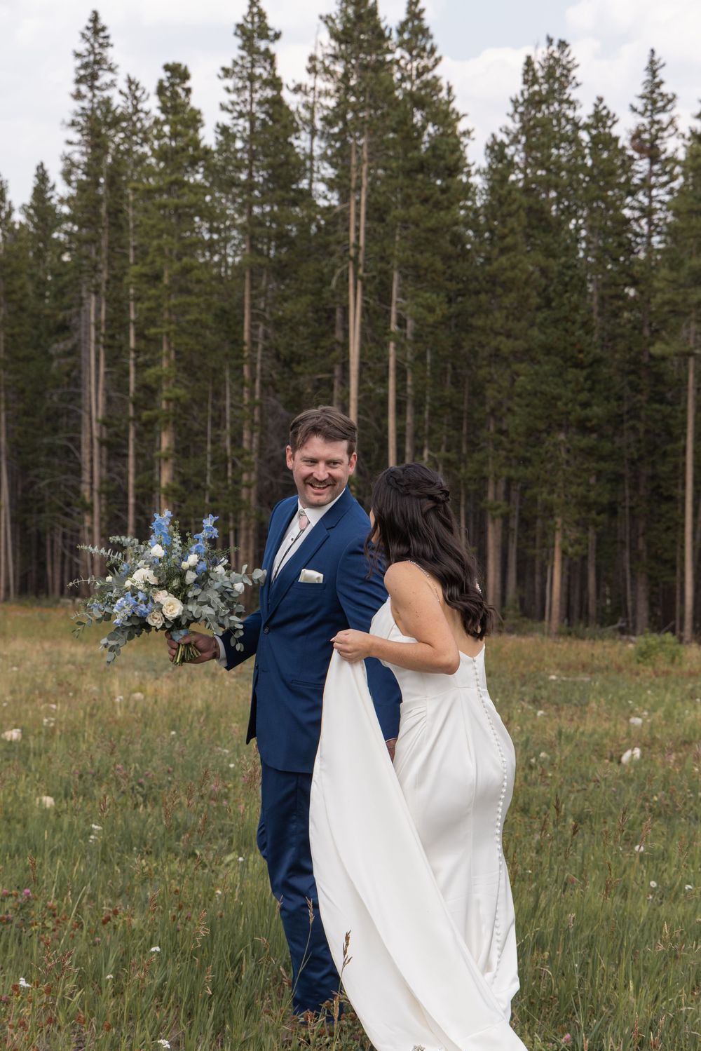 Bride in white gown and groom in navy suit share a joyful first look moment in a wildflower meadow with pine trees.