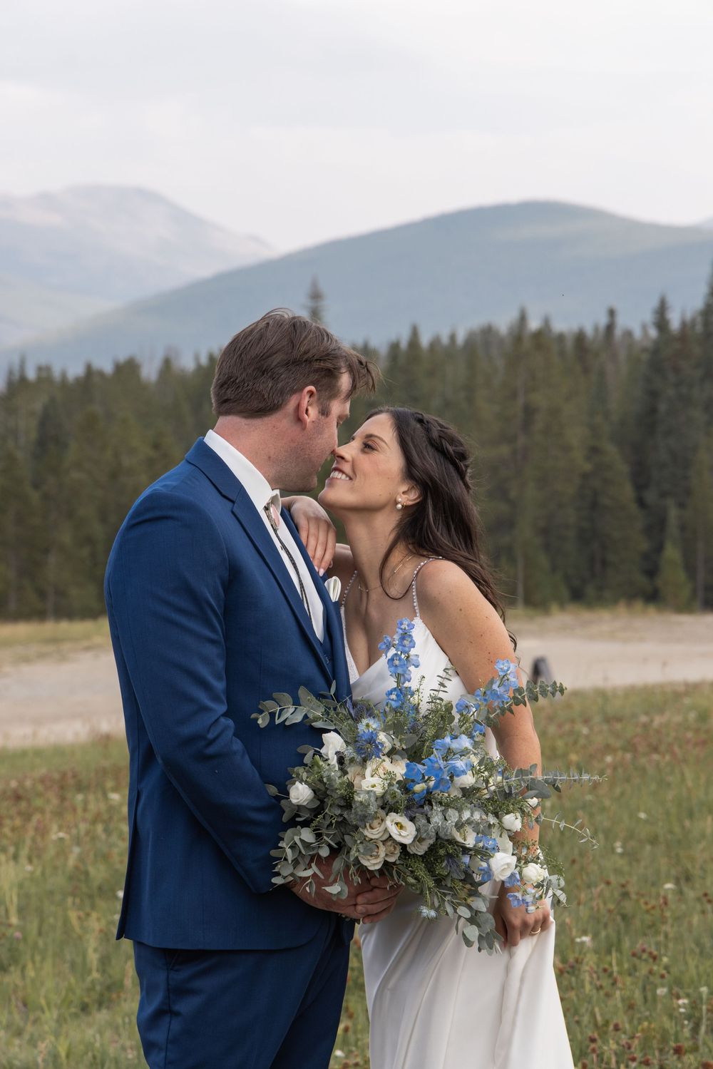 Bride and groom share a romantic moment outdoors with mountain and forest backdrop, bride holding blue and white bouquet.