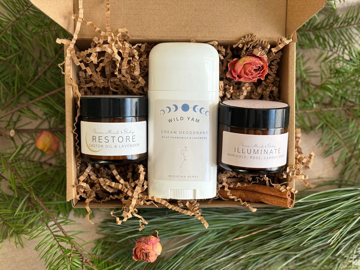 A natural skincare gift set featuring Wild Yam deodorant, Restore balm, and Illuminate cream in a kraft box with dried botanicals.
