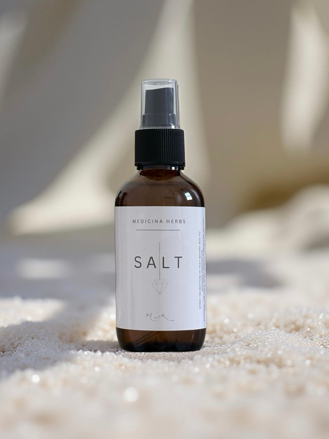 Medicina Herbs SALT spray bottle in amber glass with black pump, displayed on white sand in natural sunlight.