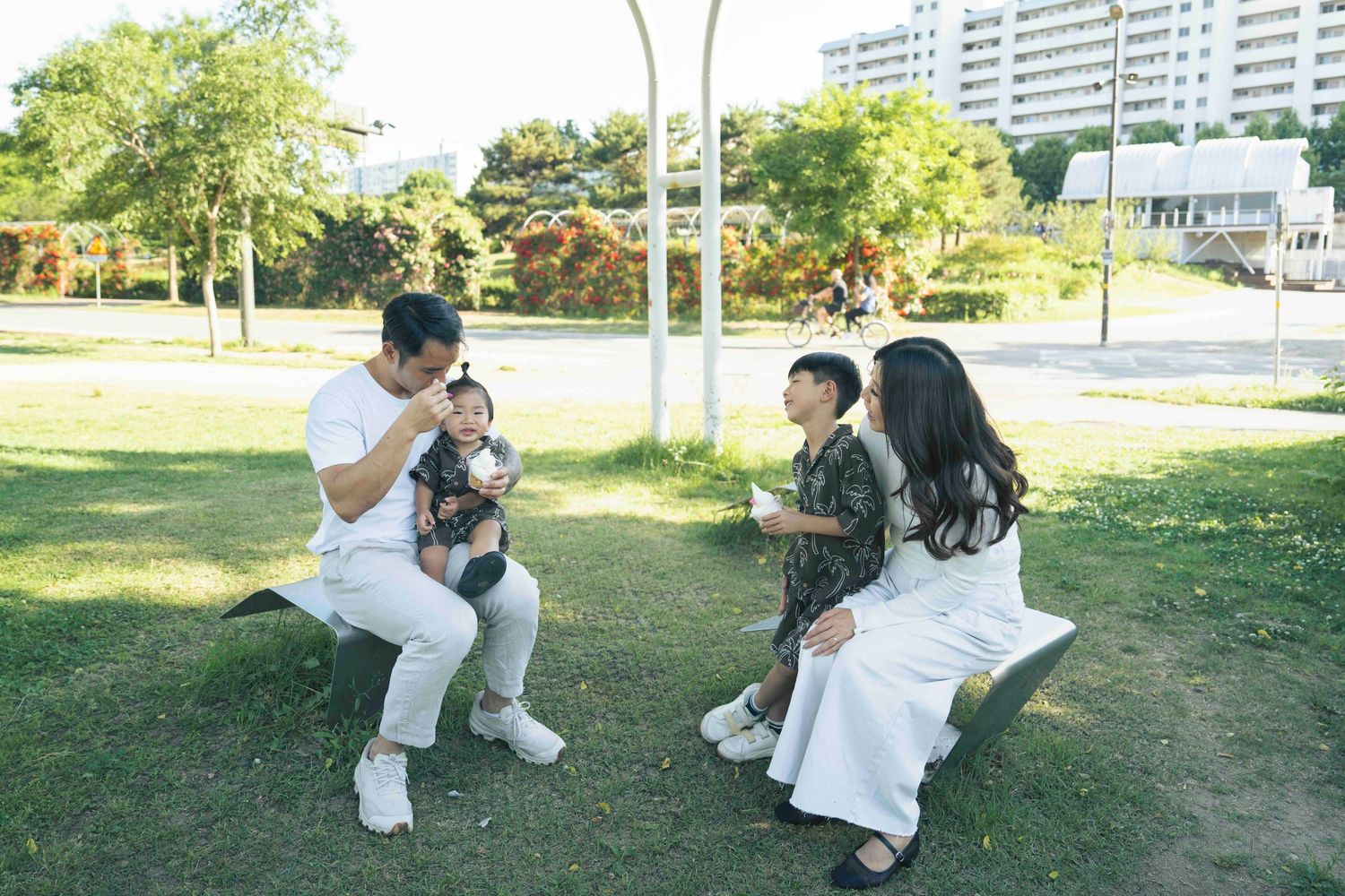 Family of four relaxing on park benches outdoors, parents holding young children on a sunny day.
