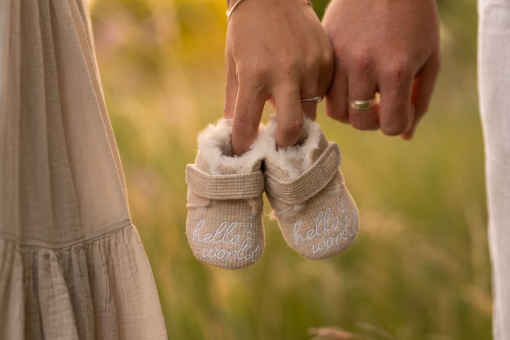 Couple holding tiny baby booties with 'hello' text outdoors, announcing pregnancy in a soft natural setting.