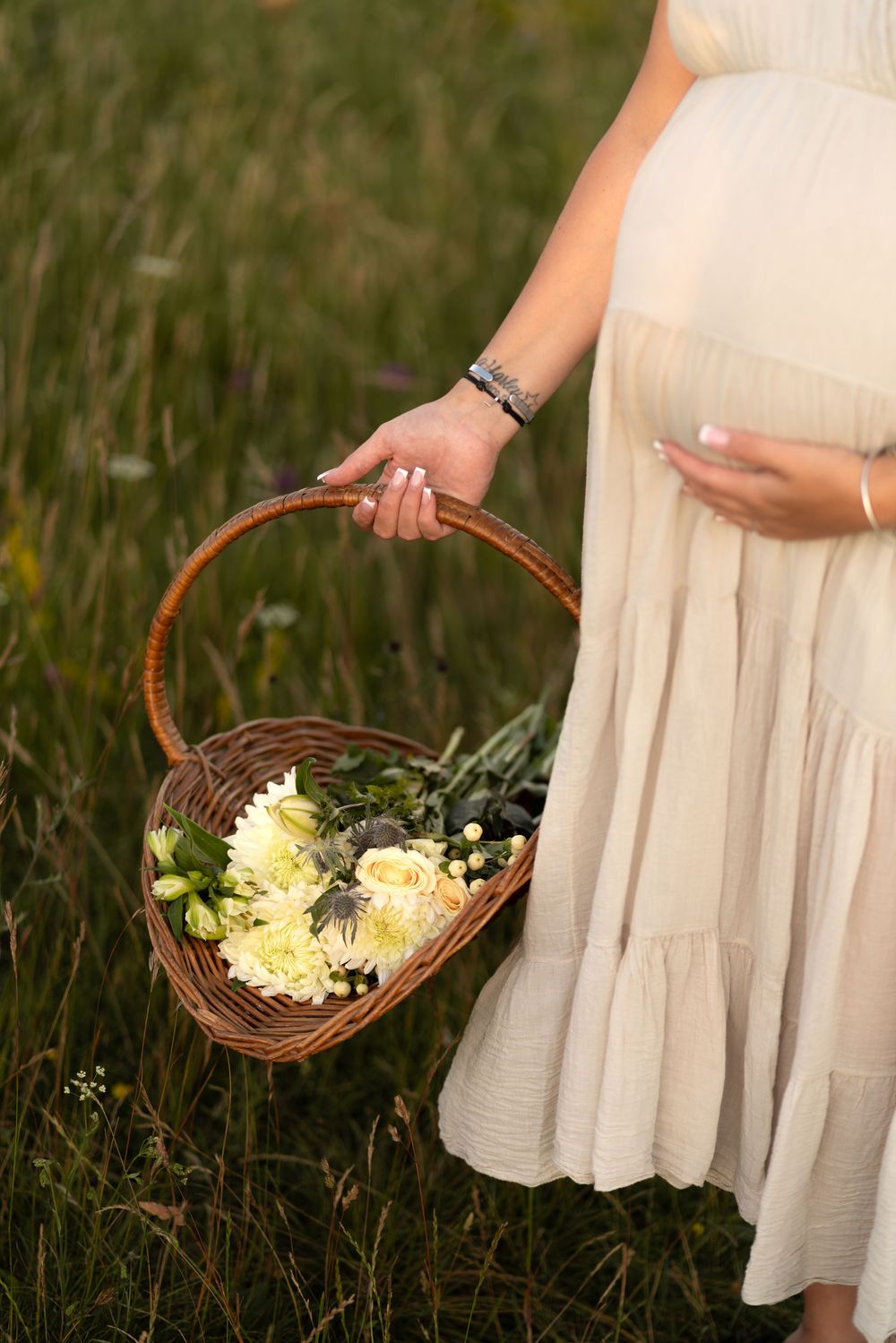 Pregnant woman in cream dress holding wicker basket with white flowers in a green meadow.