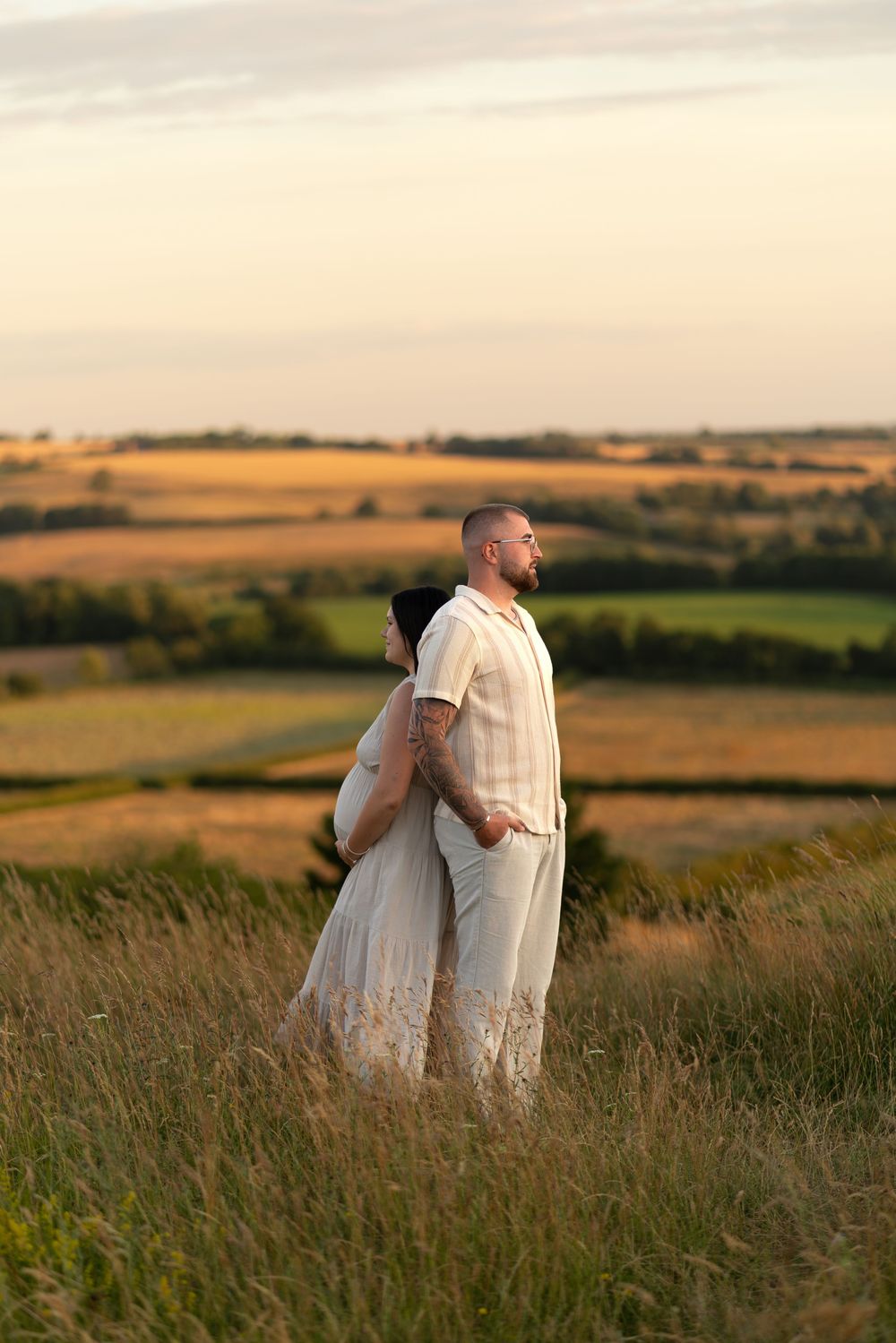 Couple in white attire embracing in a golden field at sunset with scenic countryside views.