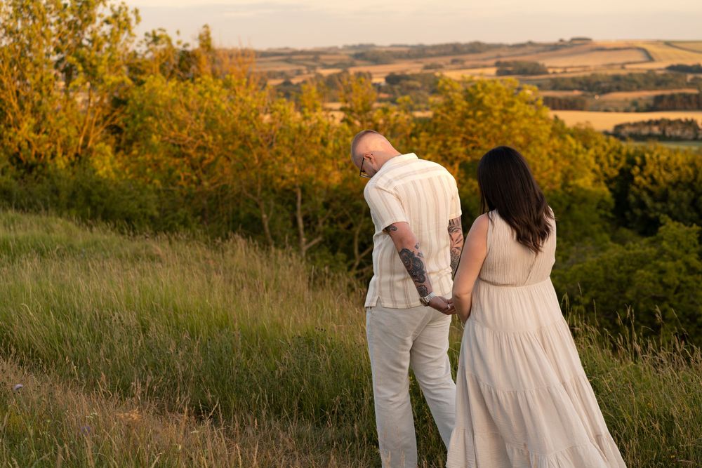 Couple holding hands walking through golden hour meadow with rolling hills and autumn trees in background.