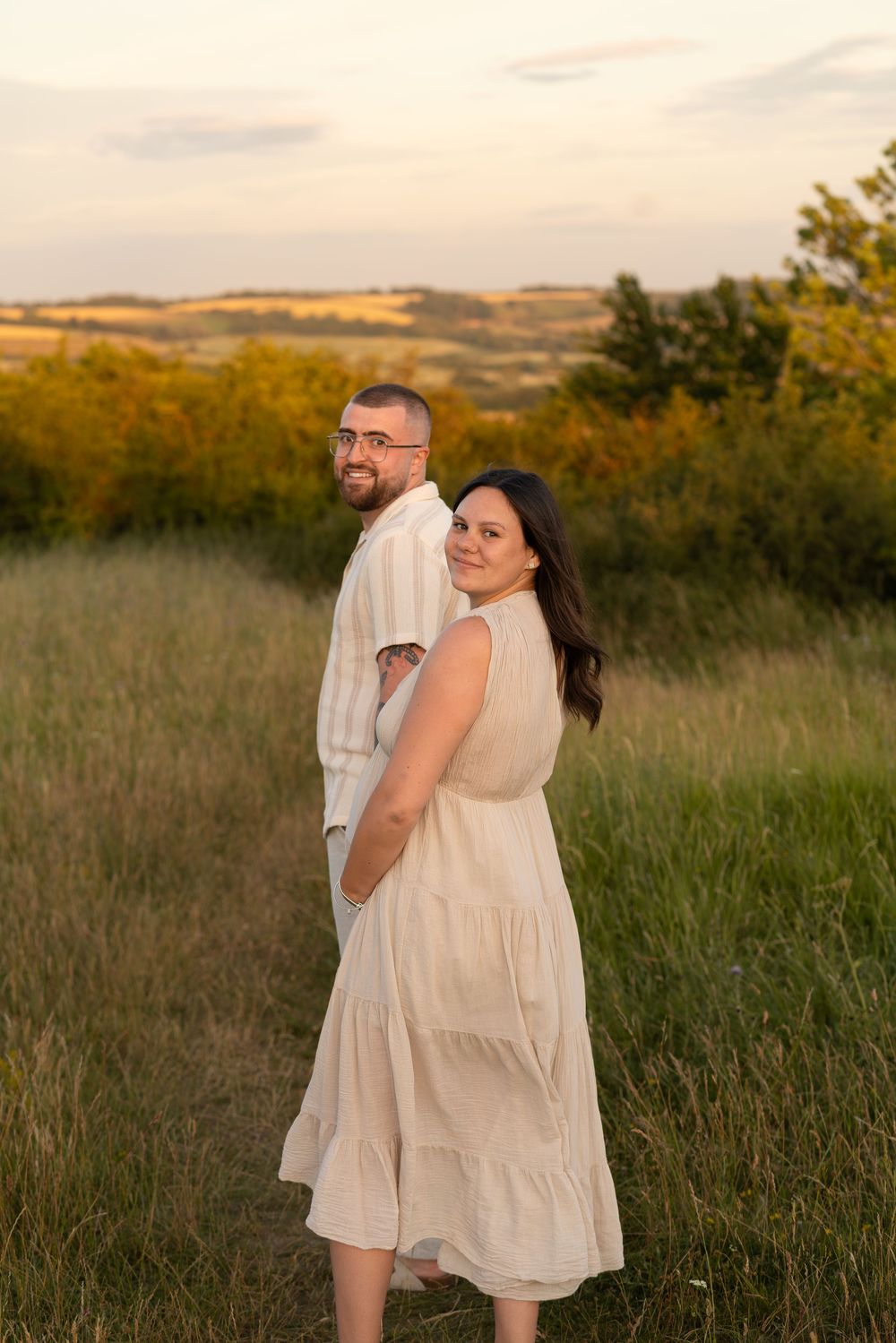 Couple in matching cream outfits posing in a golden meadow at sunset with rolling hills in the background.