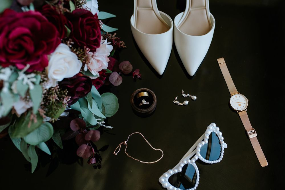 Elegant bridal flat lay with white heels, burgundy floral bouquet, gold watch, sunglasses, rings, and earrings on dark surface.