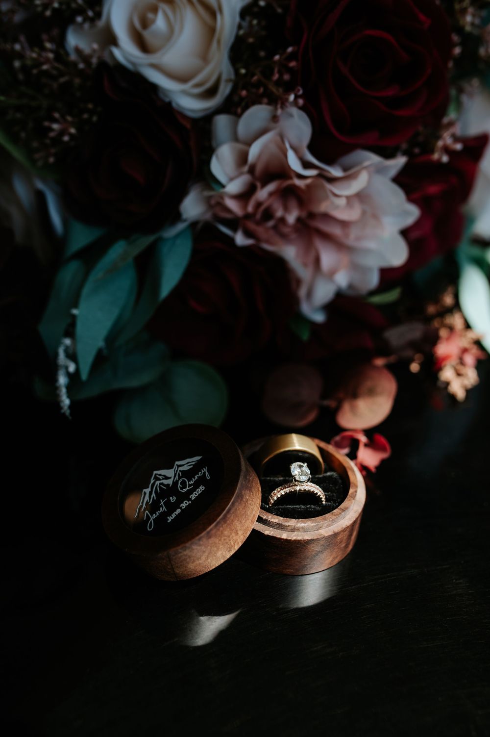 Wedding rings in a wooden box beside a bouquet of dark red, blush, and cream roses.