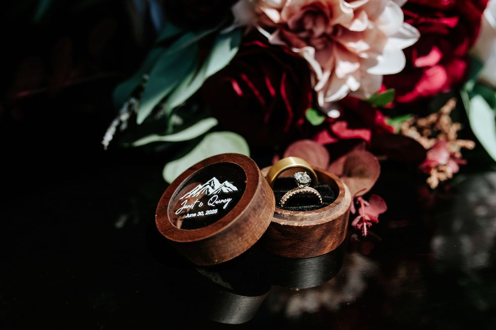 Elegant wedding rings displayed in an engraved wooden ring box surrounded by dark red and pink floral bouquet.