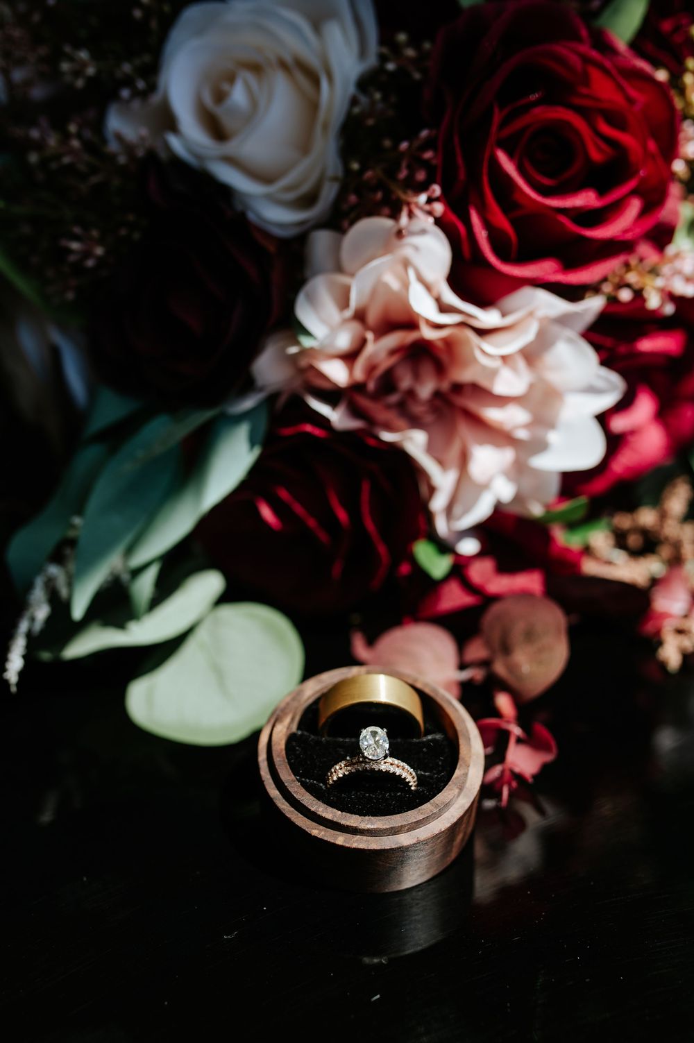 Diamond engagement ring in wooden box beside dark red and white wedding bouquet.