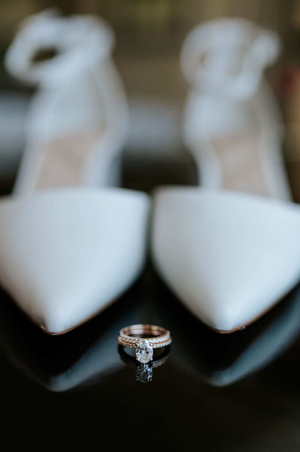 Diamond engagement ring and wedding band displayed in front of elegant white pointed-toe bridal heels on a reflective surface.