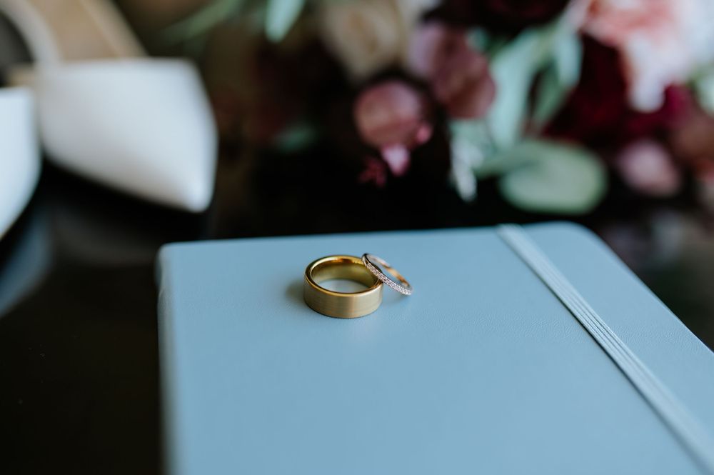 Two gold wedding bands resting on a light blue book with floral bouquet and white heels in background.