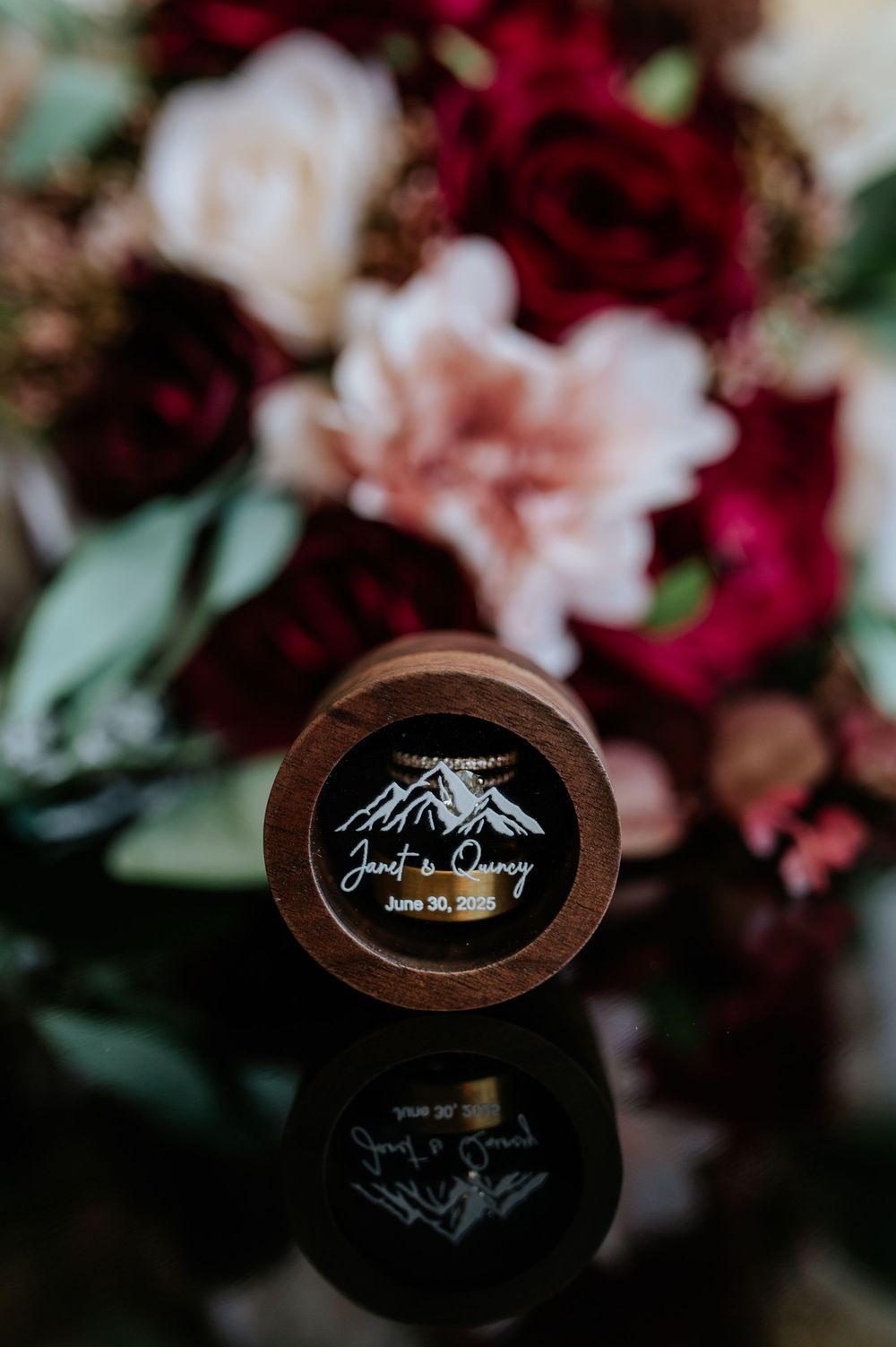 Custom wooden wedding ring box engraved with mountain design and couple names Janet & Quincy, June 30 2019, with floral backdrop.