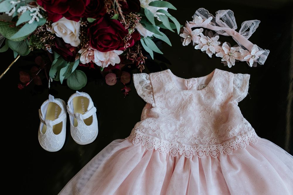 Pink baby girl baptism dress with lace details, white Mary Jane shoes, and floral headband beside red roses bouquet.
