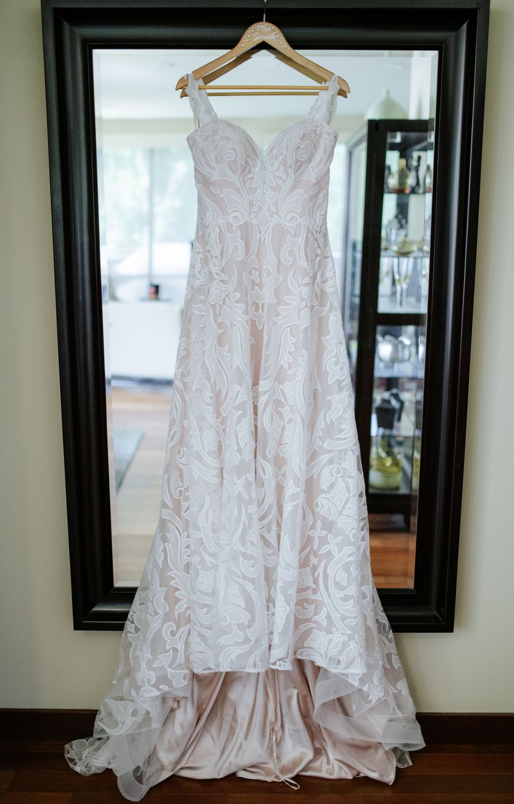 Elegant lace wedding gown with sweetheart neckline hanging on a wooden hanger in front of a large mirror.