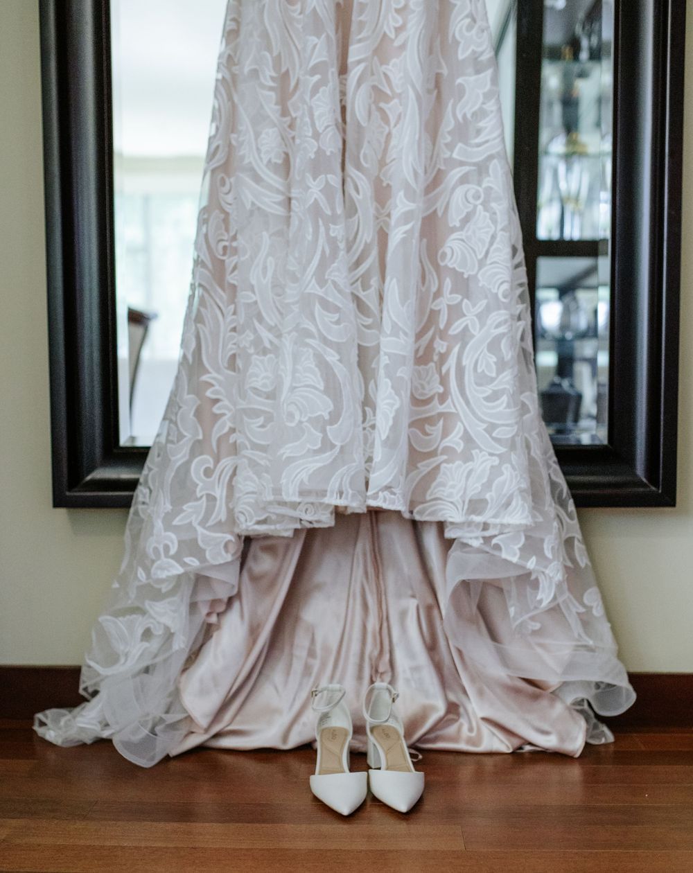 Elegant lace wedding gown with blush lining hanging near mirror with white heels below.
