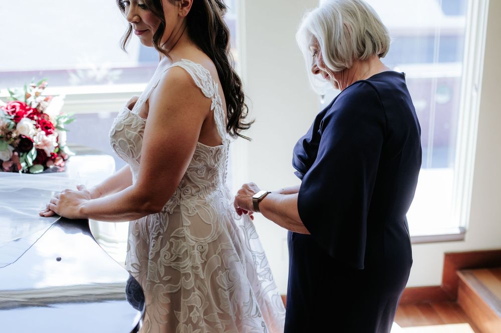 Bride in lace wedding dress being helped by older woman in navy blue outfit during wedding preparations.