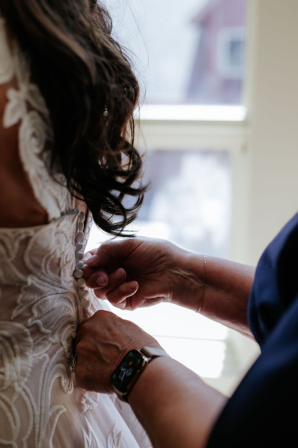 A bride in lace wedding dress gets buttons fastened by helper near bright window on wedding day.