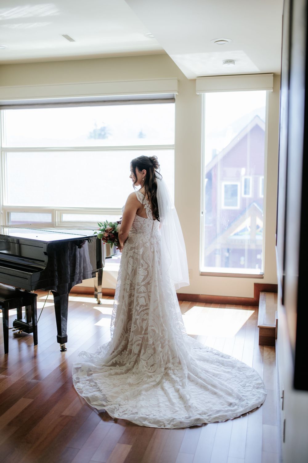 Bride in lace wedding gown with long train and veil stands by sunlit windows near a grand piano.