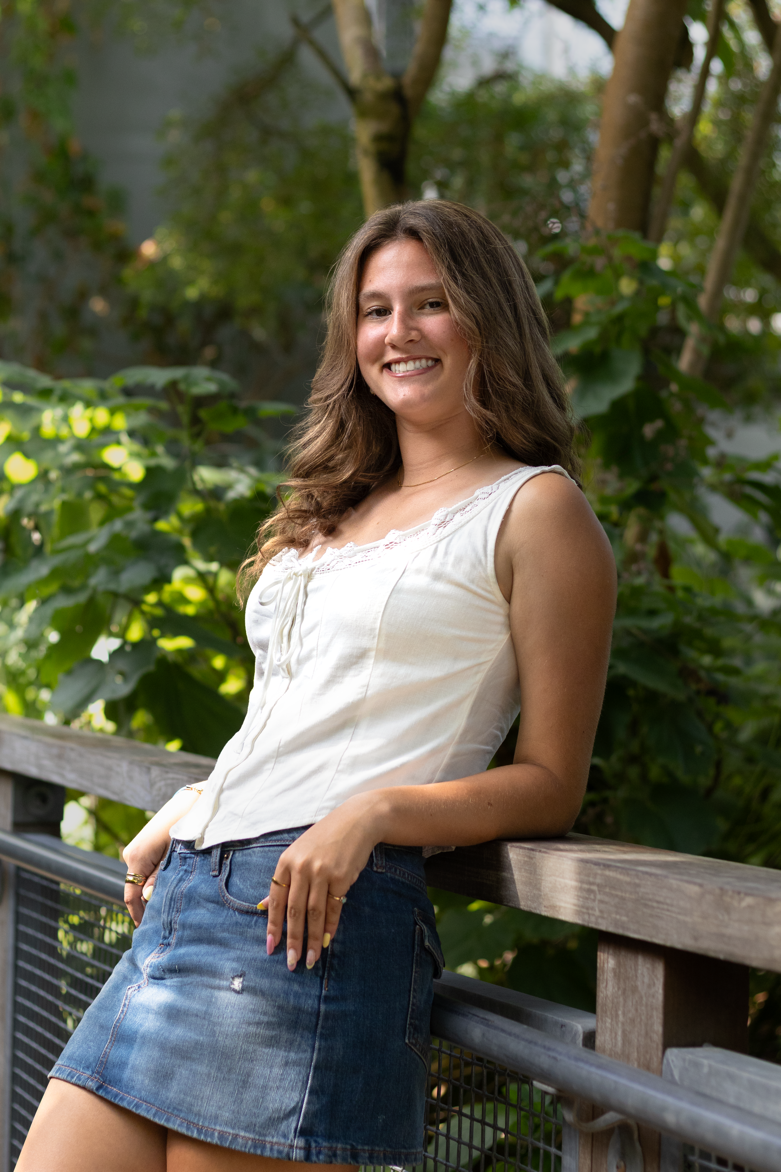 Smiling young woman in white top and denim skirt leaning on wooden railing with lush green foliage behind her.