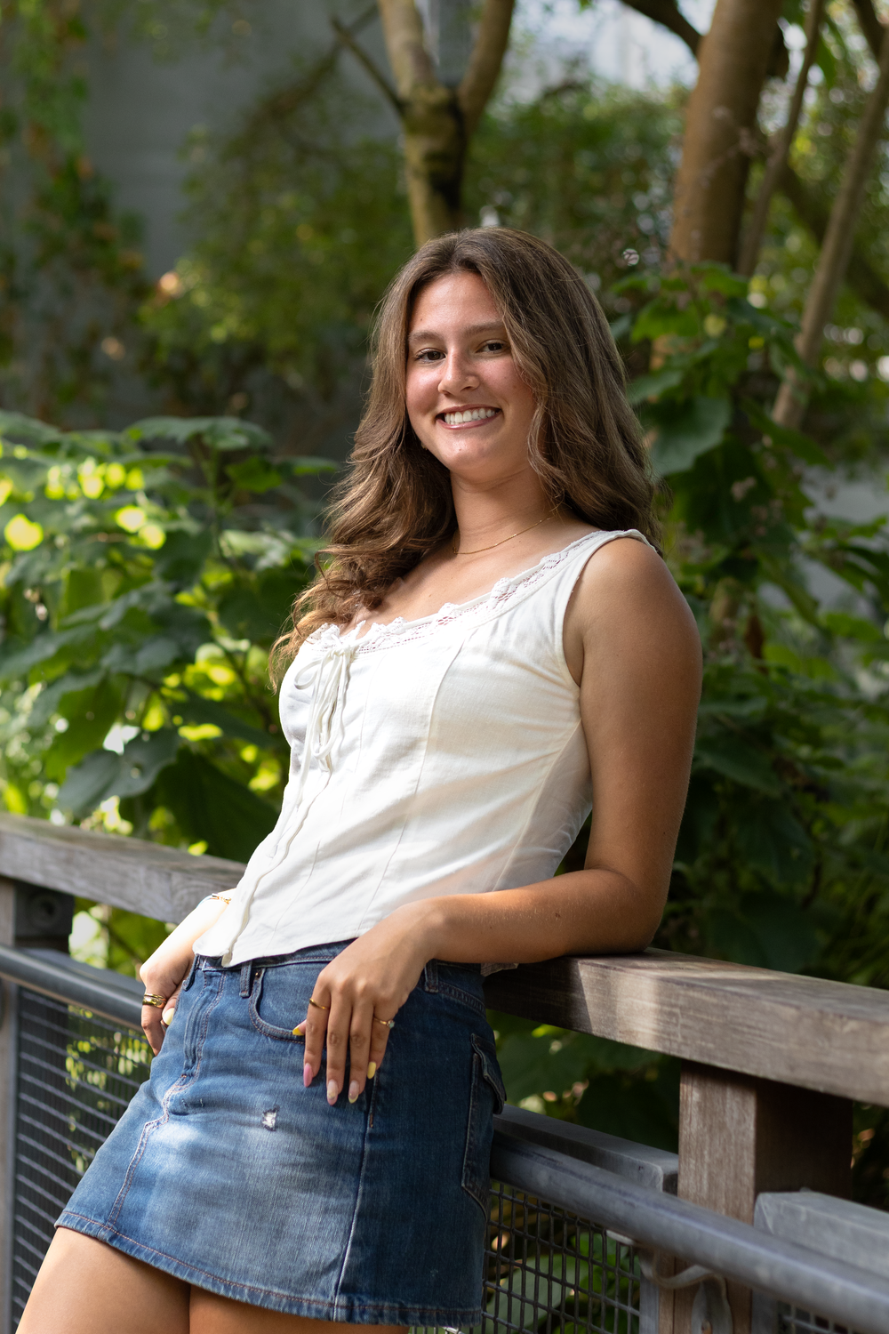 Smiling young woman in white top and denim skirt leaning on wooden railing with lush green foliage behind her.