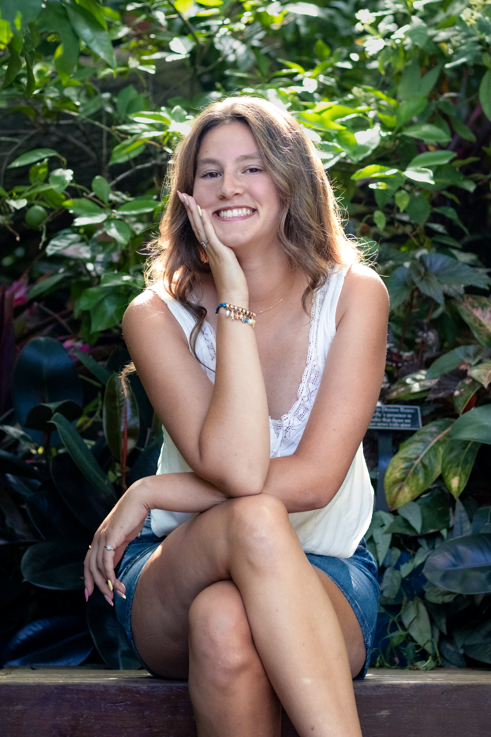 Smiling young woman in white tank top and denim shorts sitting outdoors among lush green tropical plants.