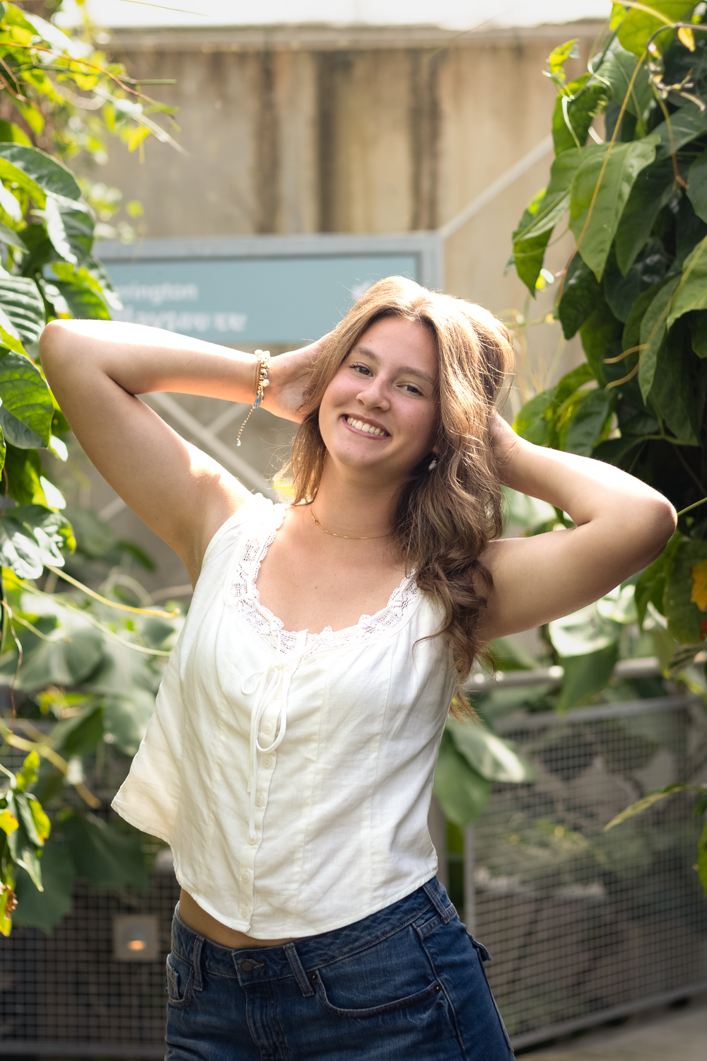 Smiling young woman with long hair poses outdoors in white top and jeans, surrounded by lush green tropical plants.