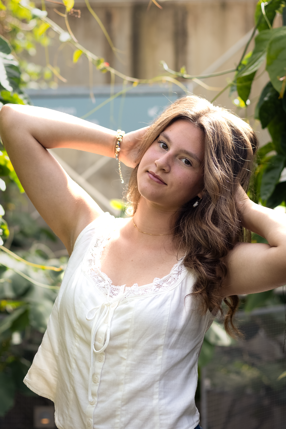 Young woman with long brown hair poses outdoors in white lace top surrounded by lush green vines.