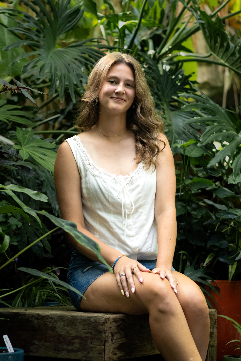Young woman with wavy blonde hair sits smiling in lush tropical garden wearing white top and denim shorts.