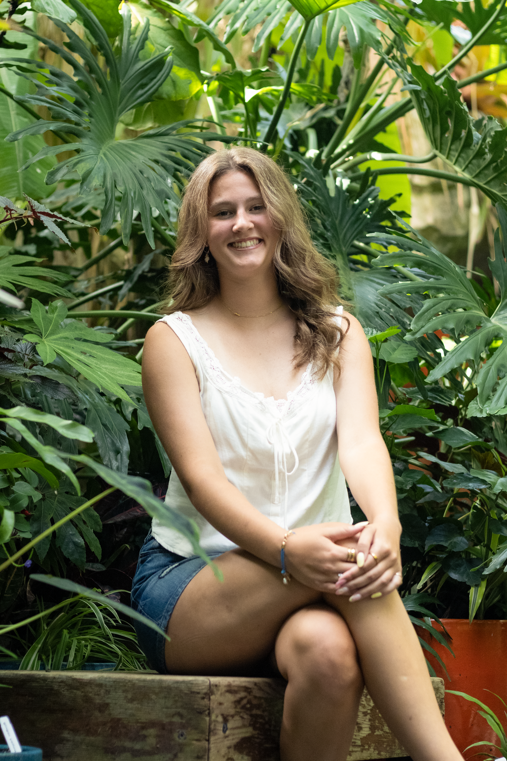 Young woman smiling in white top and denim shorts, seated among lush tropical green plants in a garden setting.