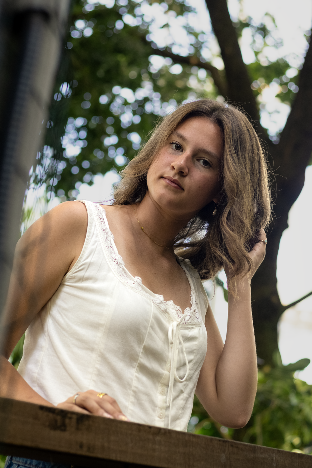 Young woman in white lace tank top poses outdoors near wooden railing with green trees in background.