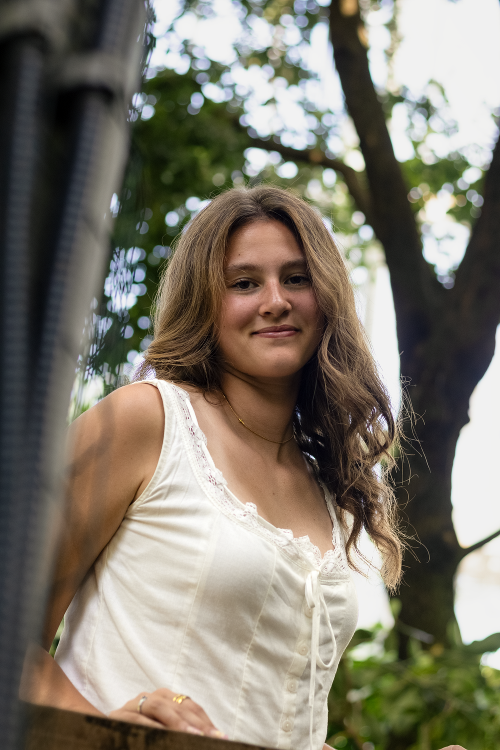Young woman with long wavy hair smiling in a white sleeveless top, posed outdoors with trees in background.