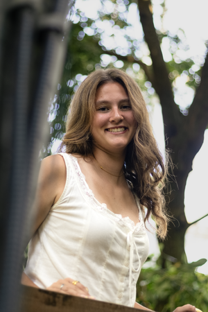 Smiling young woman with long brown hair wearing a white lace top, photographed outdoors with trees in background.