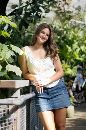 Young woman in white top and denim mini skirt smiling while leaning on a railing in a lush greenhouse.