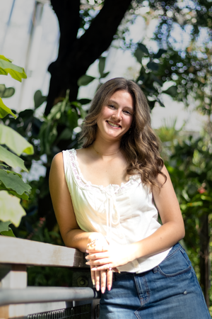 Smiling young woman with wavy brown hair in white top and jeans posing outdoors among lush green foliage.