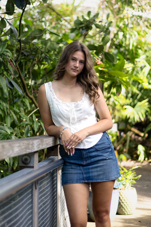 Young woman in white lace top and denim mini skirt posing by a railing in a lush tropical garden.
