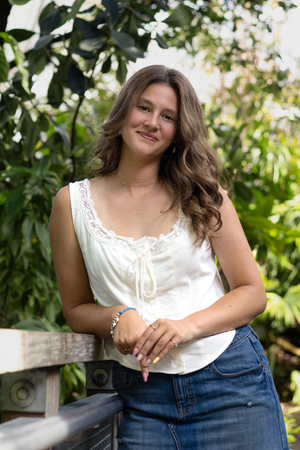 Young woman in white lace top and jeans smiling outdoors among lush green tropical plants.