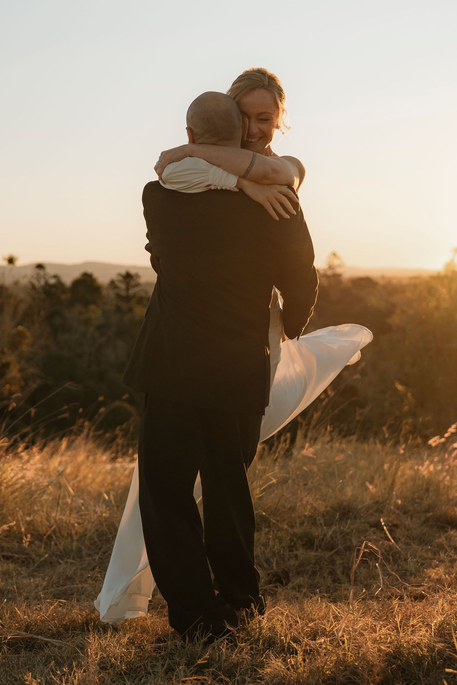 A couple embraces at golden hour in a field, bride in white gown lifted by groom in black suit during sunset wedding.