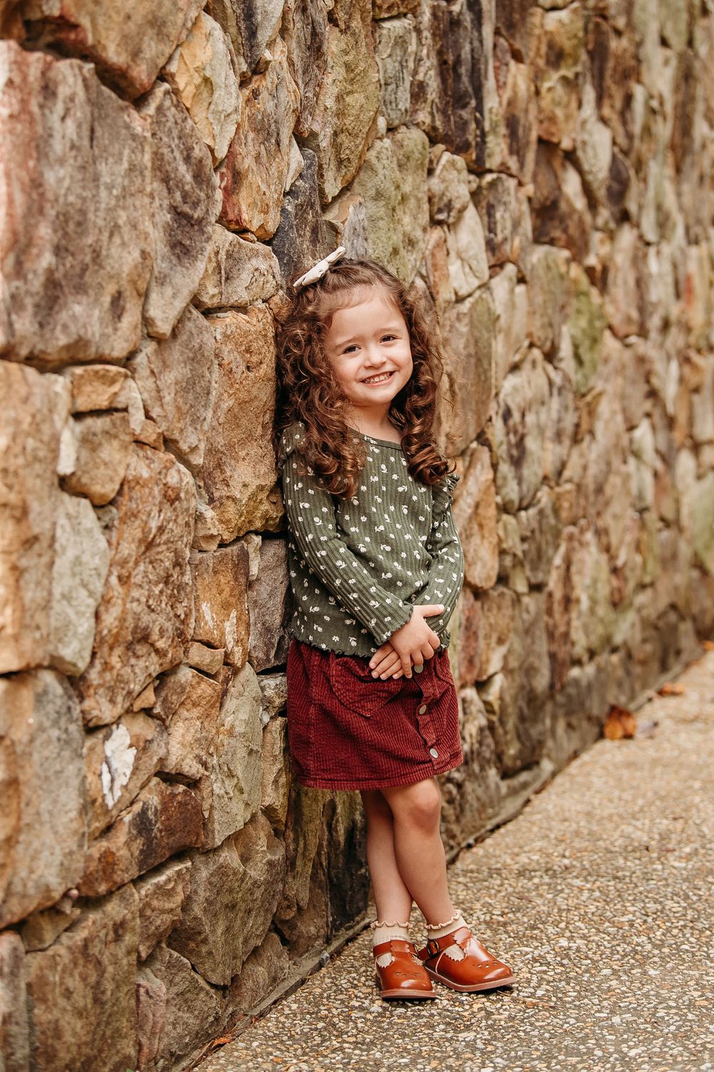 Adorable curly-haired girl in green top and red skirt leaning against a stone wall.