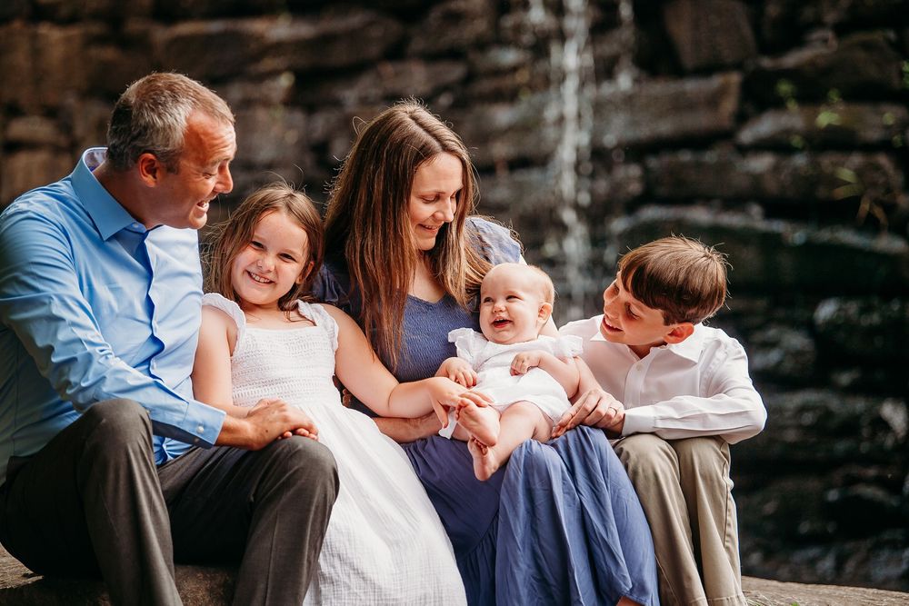 Family of five sitting together near a waterfall, smiling and bonding during outdoor portrait session.