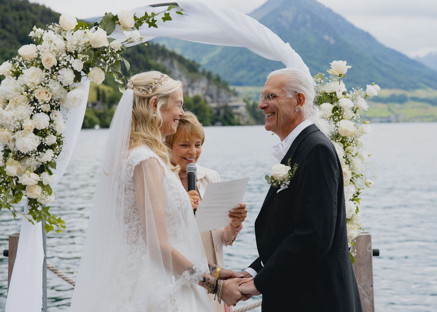 Bride and groom exchanging vows outdoors by a lake with mountain views and white floral arch.