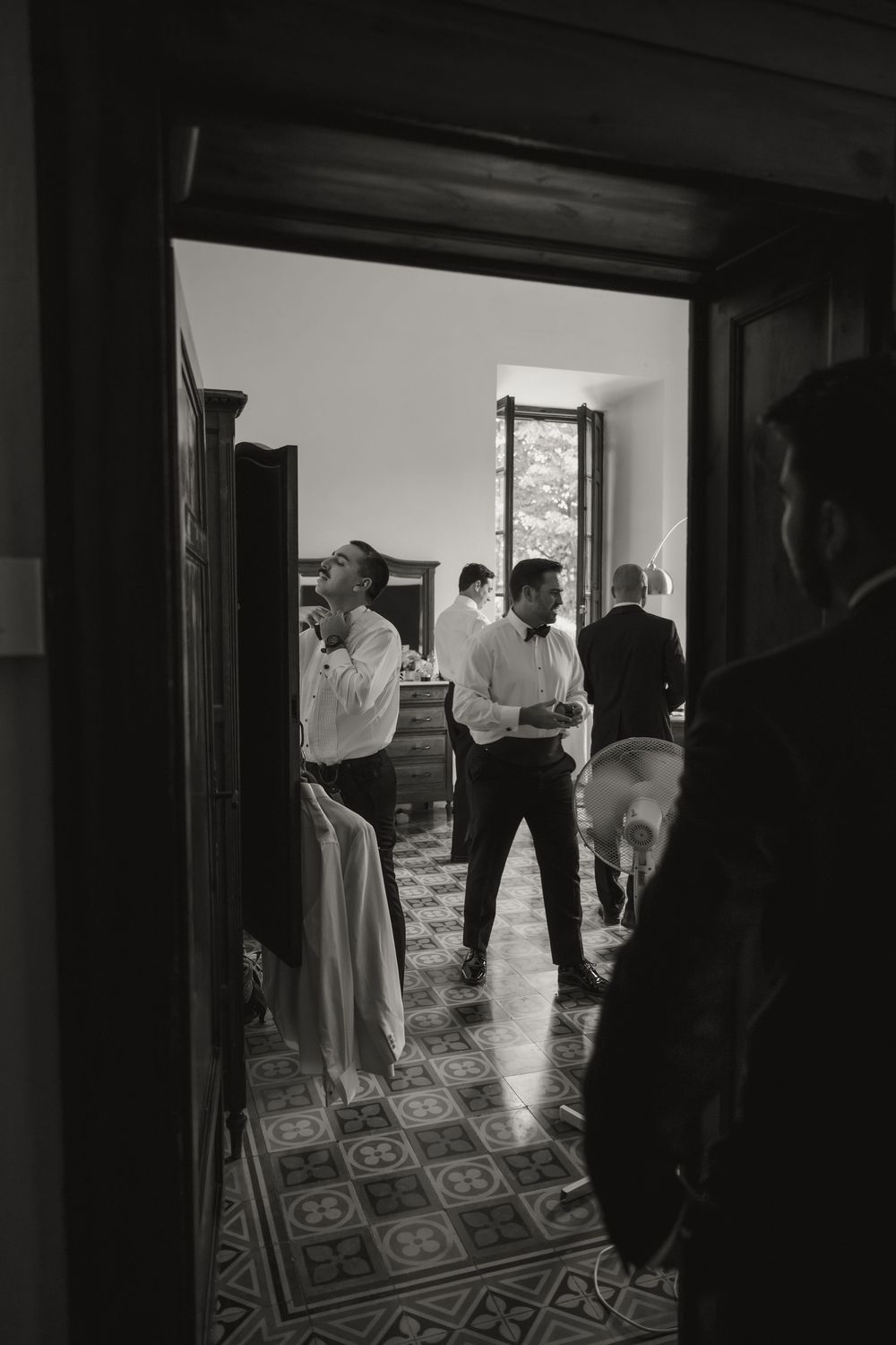 Black and white photo of groomsmen getting ready in elegant room with patterned tile floor before wedding ceremony.