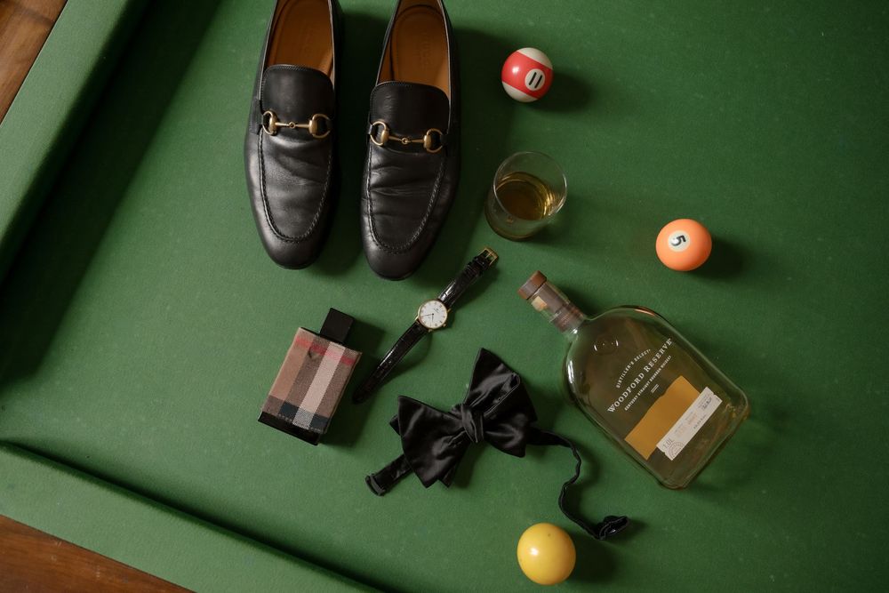 Flat lay of mens accessories on green felt including Gucci loafers, bow tie, watch, whiskey bottle, and billiard balls.