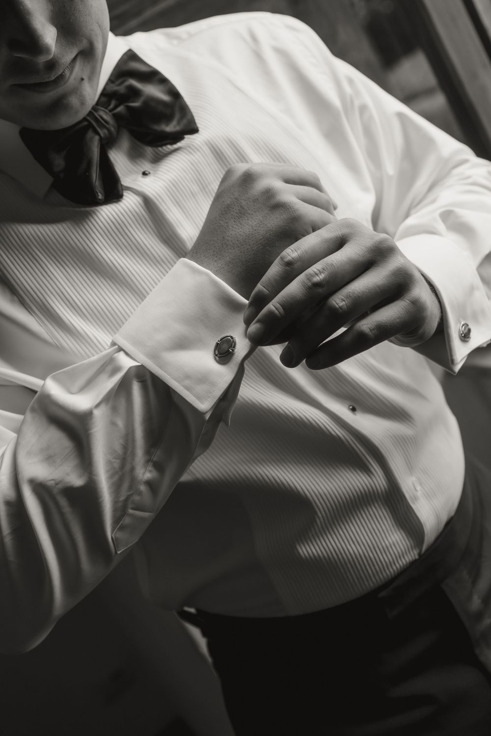Black and white photo of a groom in a tuxedo shirt and bow tie fastening his cufflinks.