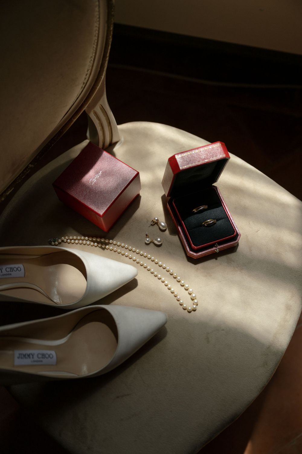 Bridal accessories on chair: Jimmy Choo heels, pearl necklace, earrings, and wedding rings in red Cartier box.