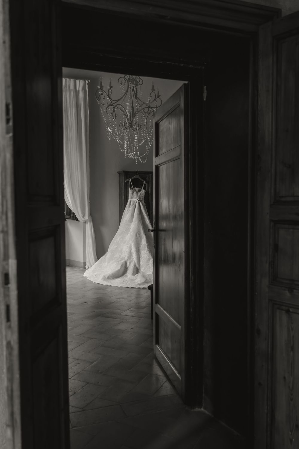 Black and white photo of an elegant wedding dress hanging in a sunlit room viewed through an open doorway with chandelier.