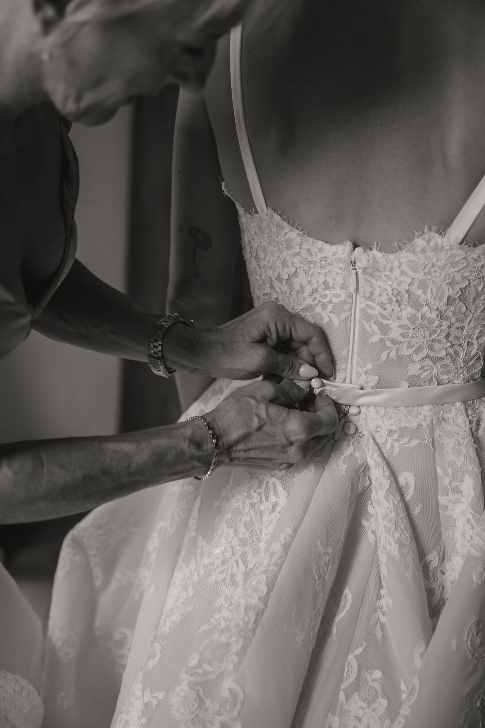 Black and white photo of hands fastening a bow on the back of a lace wedding dress.