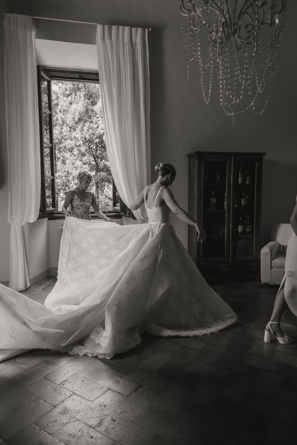 Black and white photo of a bride in an elegant lace gown with a long train in a classic chandelier-lit room.