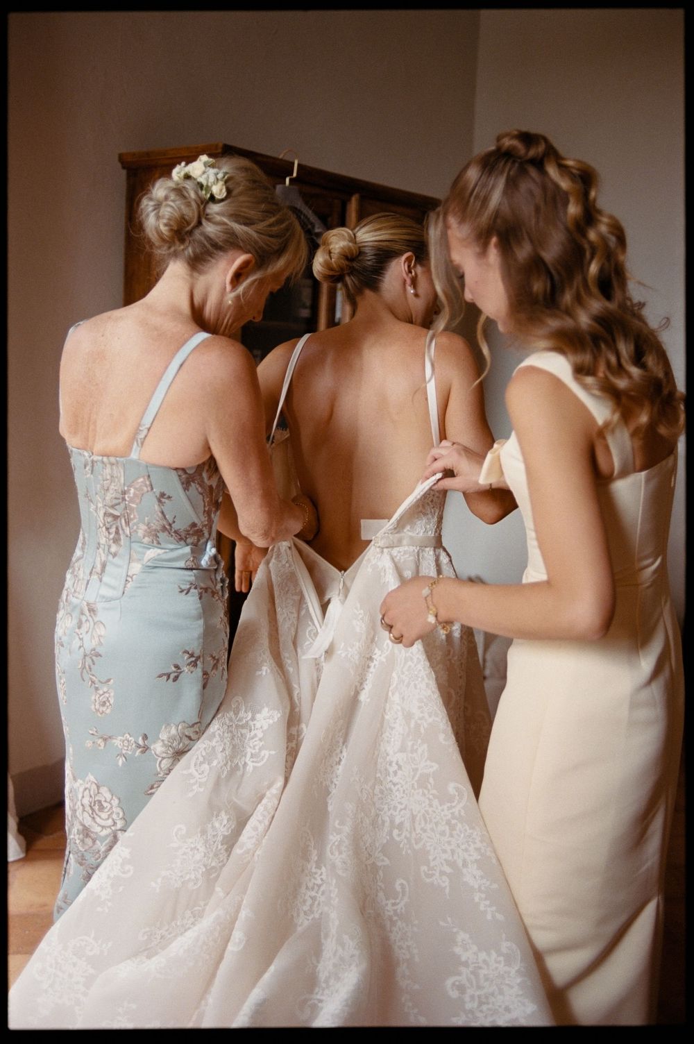 Two bridesmaids help a bride into her lace wedding gown from behind, preparing for the ceremony.