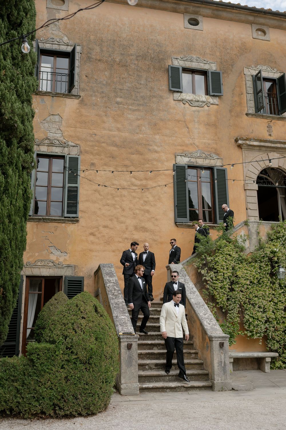 Groomsmen in tuxedos descend stone steps of a rustic Italian villa with green shutters and string lights.